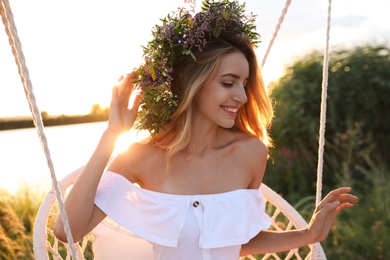 Photo of Young woman wearing wreath made of beautiful flowers on swing chair outdoors at sunset