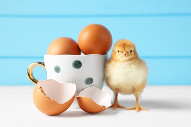 Photo of Cute chick with cup, eggs and pieces of shell on white wooden table, closeup. Baby animal