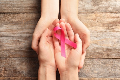 Women holding pink ribbon on wooden background, top view. Cancer awareness