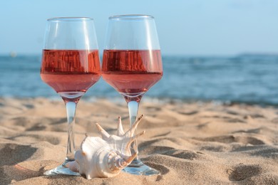Photo of Glasses of tasty rose wine and seashell on sand near sea, space for text