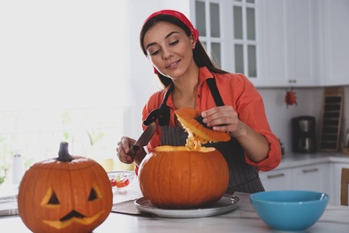 Photo of Woman making pumpkin jack o'lantern at table in kitchen. Halloween celebration