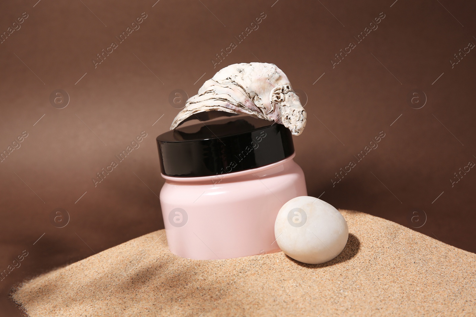 Photo of Jar of body cream, seashell and stone on sand against brown background