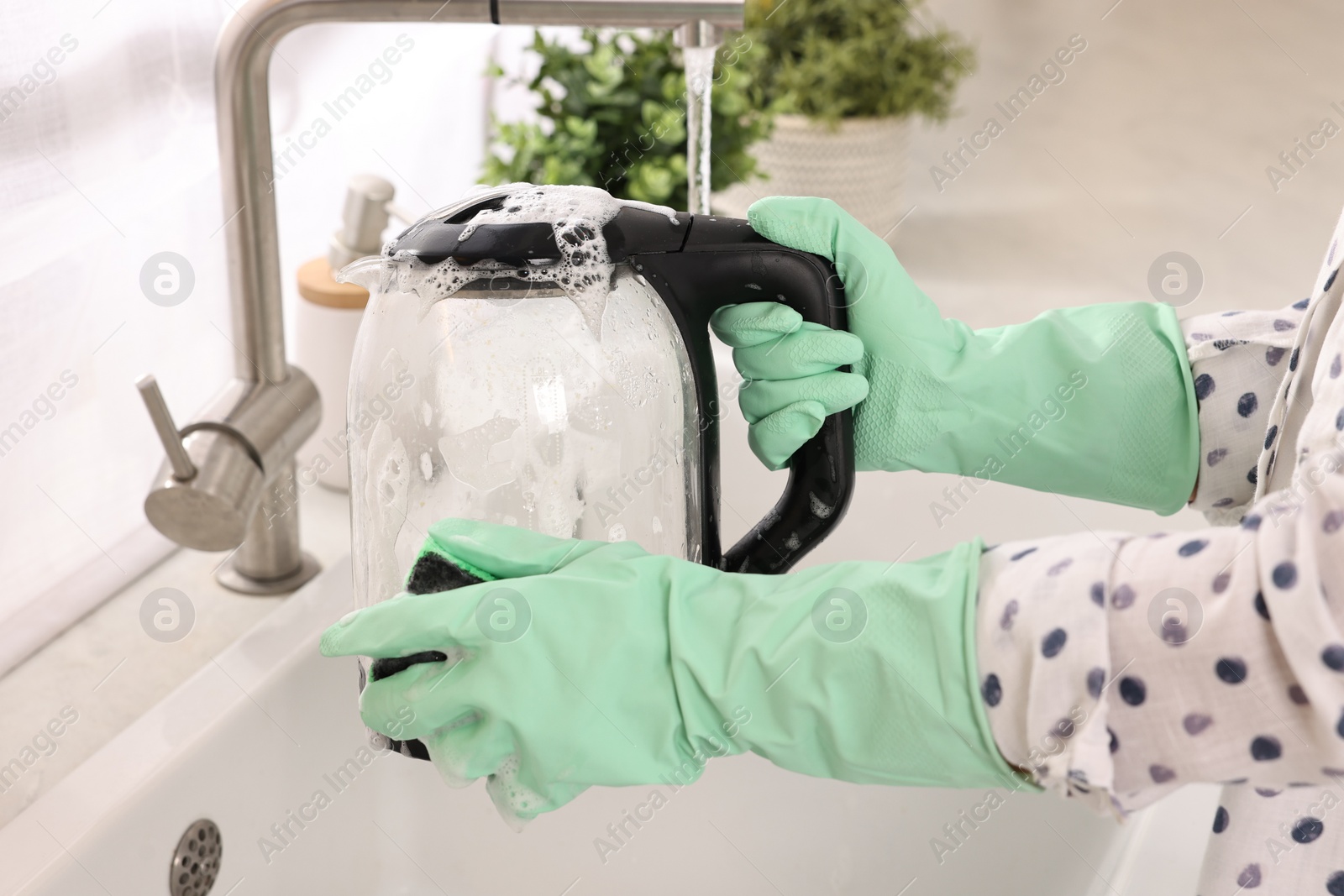 Photo of Woman washing electric kettle with sponge at sink in kitchen, closeup