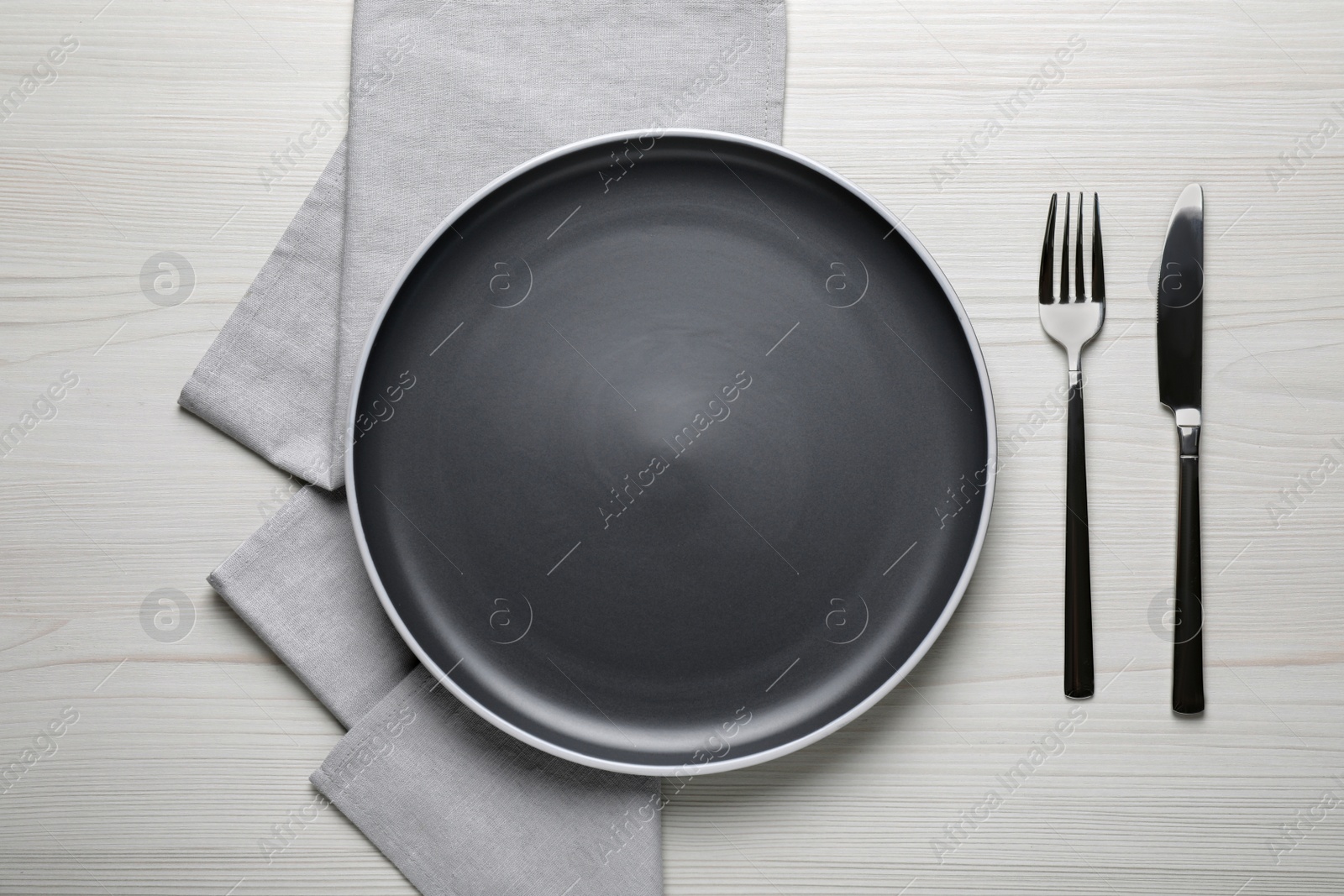 Photo of New dark plate and cutlery on white wooden table, flat lay