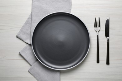 Photo of New dark plate and cutlery on white wooden table, flat lay