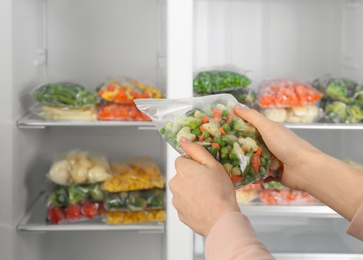 Photo of Woman holding plastic bag with frozen vegetables near open refrigerator, closeup