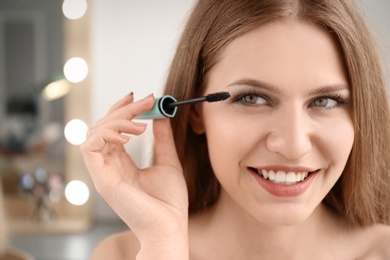 Photo of Attractive young woman applying mascara on her eyelashes against blurred background