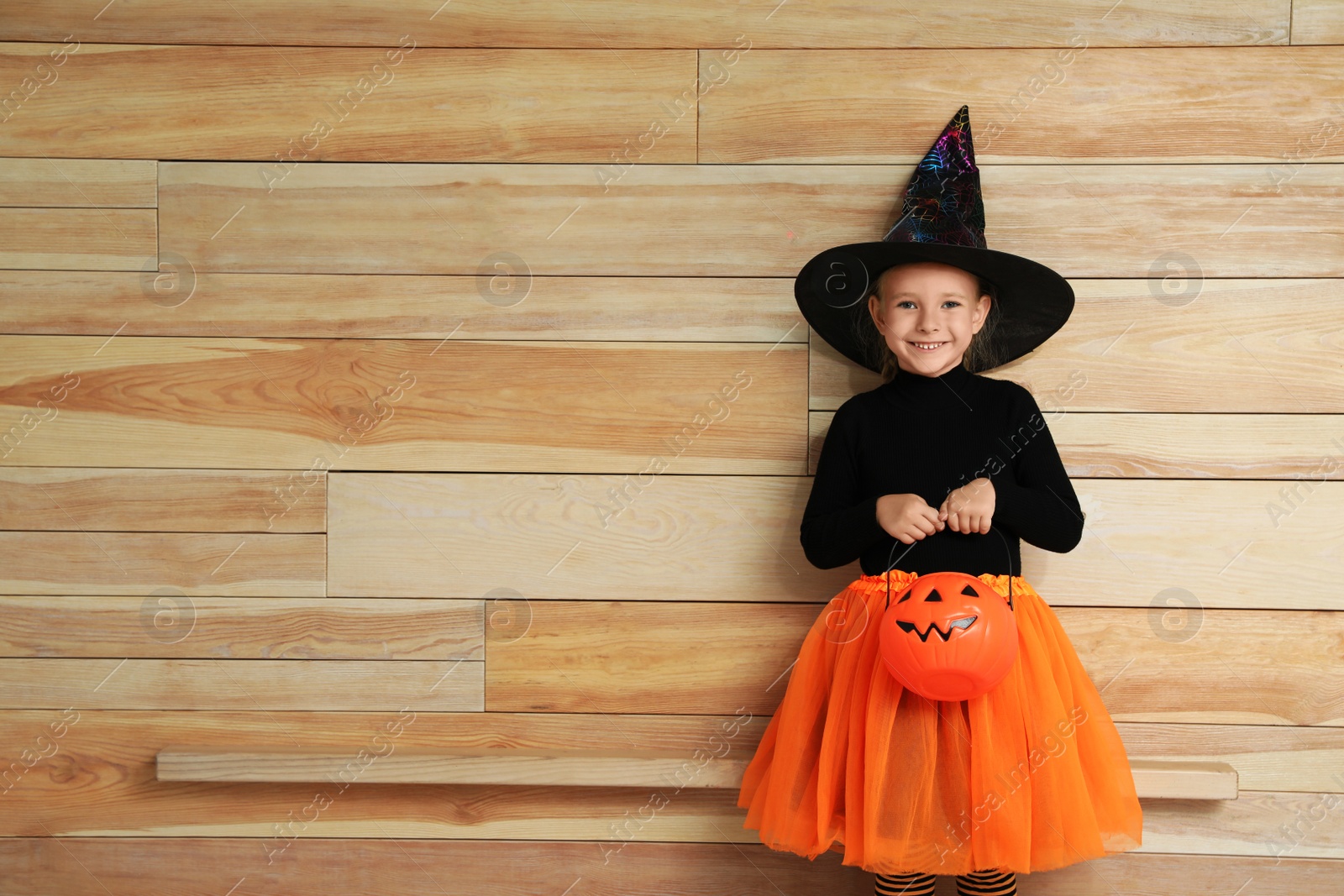 Photo of Cute little girl with pumpkin candy bucket wearing Halloween costume on wooden background. Space for text