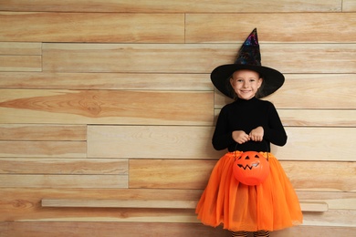 Photo of Cute little girl with pumpkin candy bucket wearing Halloween costume on wooden background. Space for text