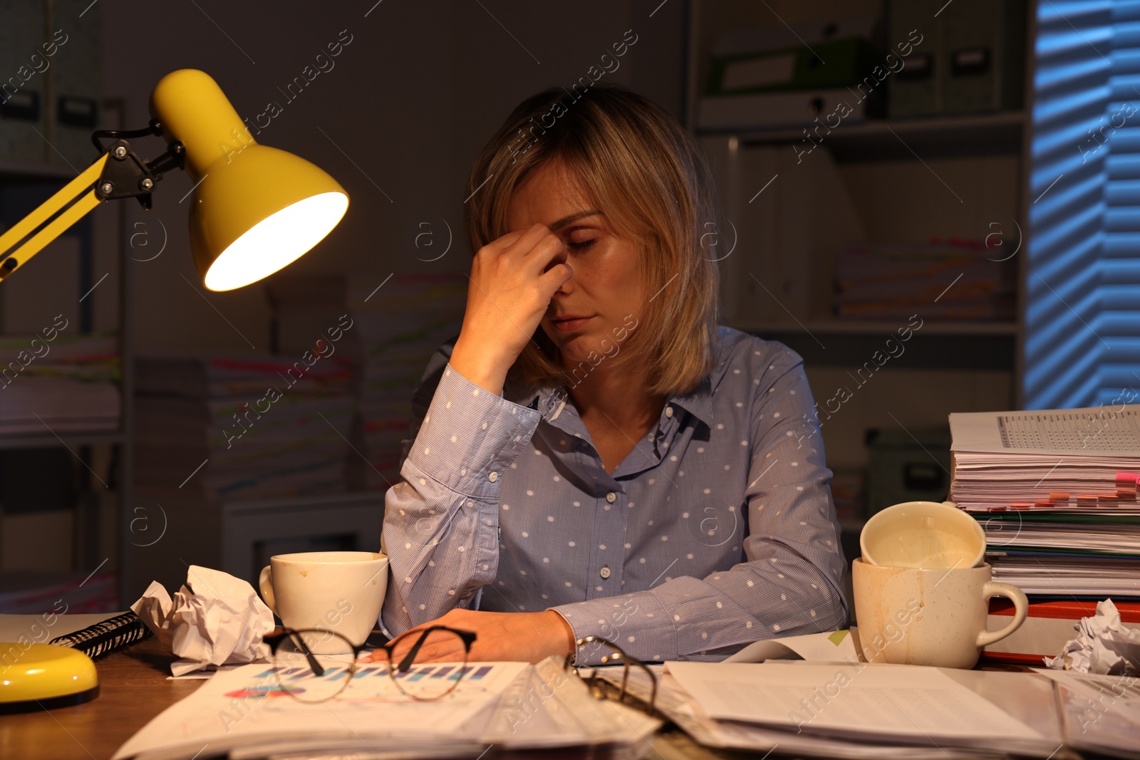 Photo of Overwhelmed woman surrounded by documents and dirty cups at table in office at night