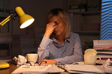 Overwhelmed woman surrounded by documents and dirty cups at table in office at night