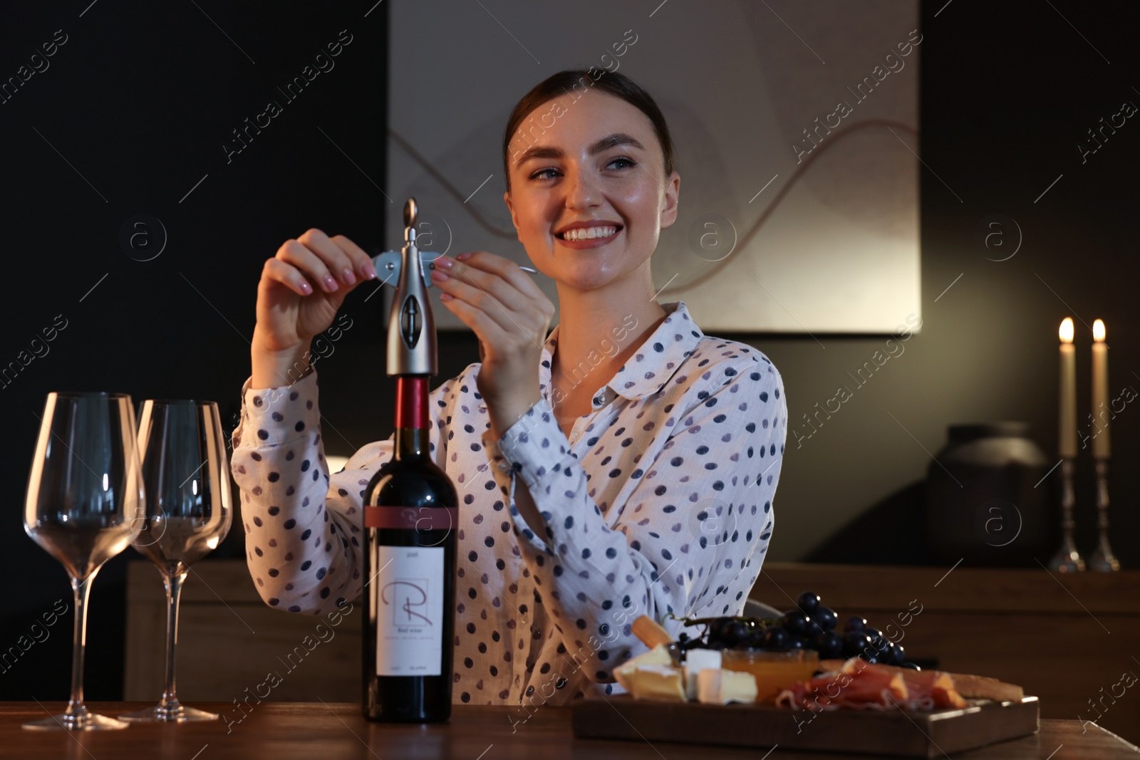 Photo of Romantic dinner. Happy woman opening wine bottle with corkscrew at table indoors