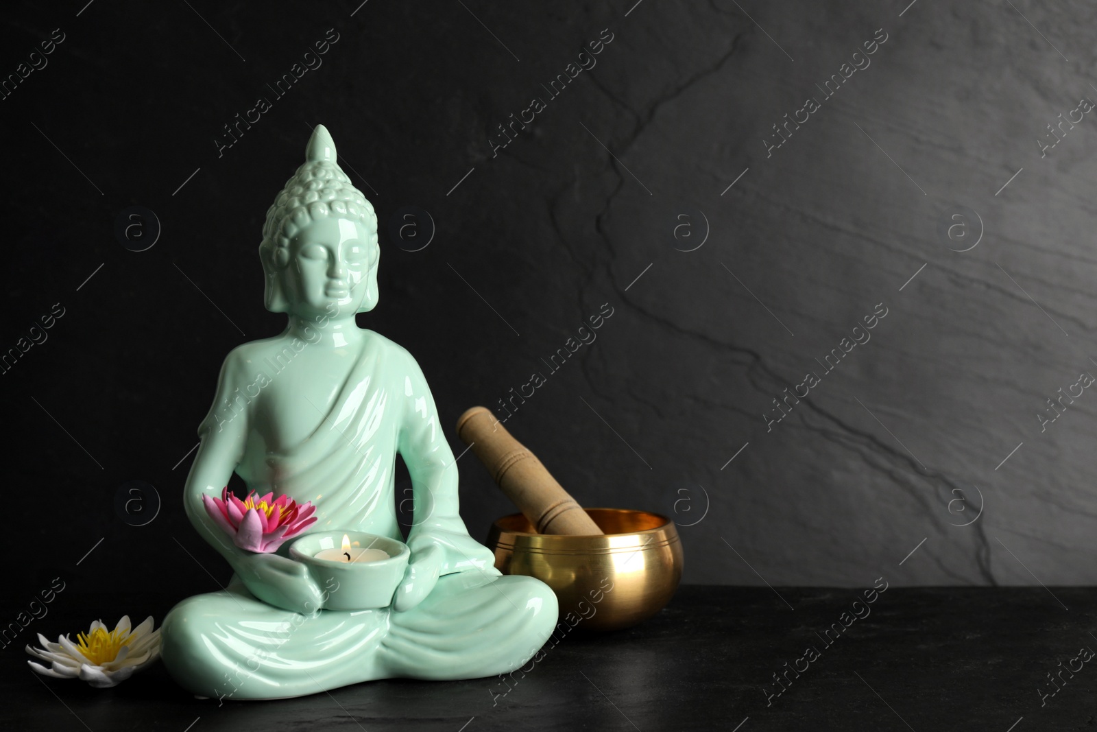 Photo of Buddha statue with burning candle, lotus flowers and singing bowl on black table. Space for text