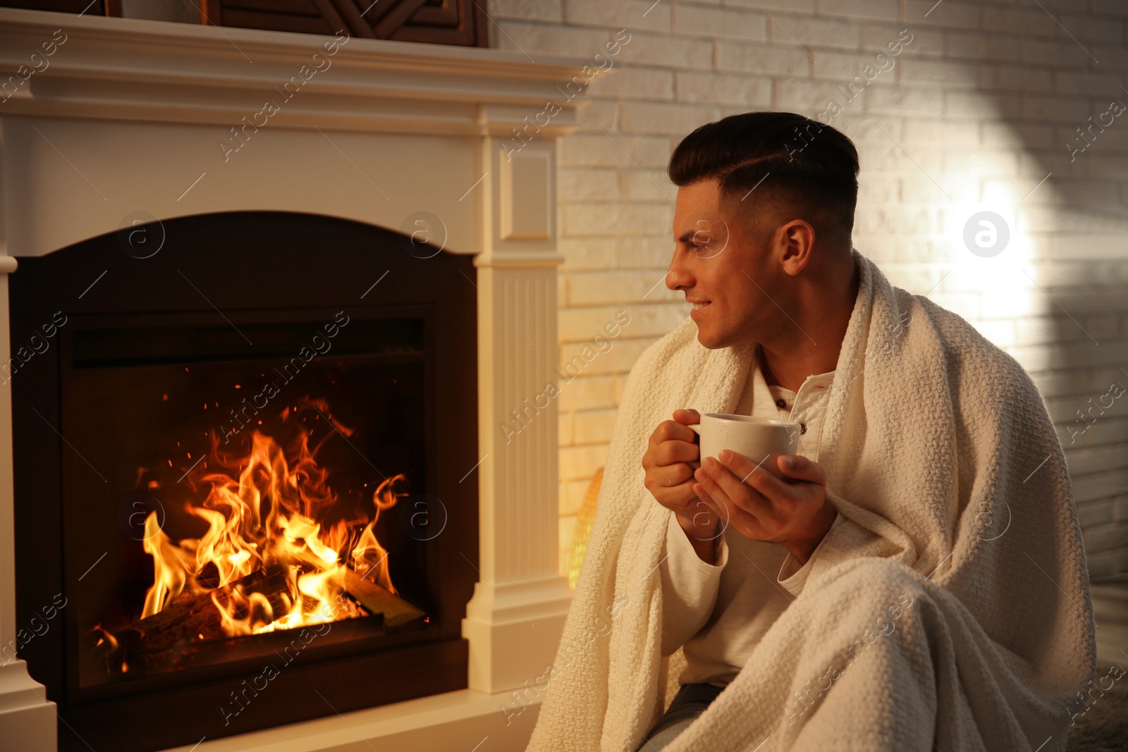 Photo of Man covered with white plaid enjoying cup of tea near fireplace