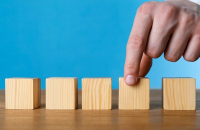 Photo of Man arranging empty cubes in row on wooden table against light blue background, closeup. Space for text