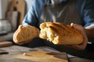 Photo of Man breaking loaf of fresh bread at wooden table indoors, closeup