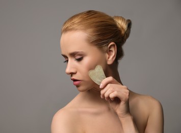 Beautiful young woman doing facial massage with gua sha tool on grey background