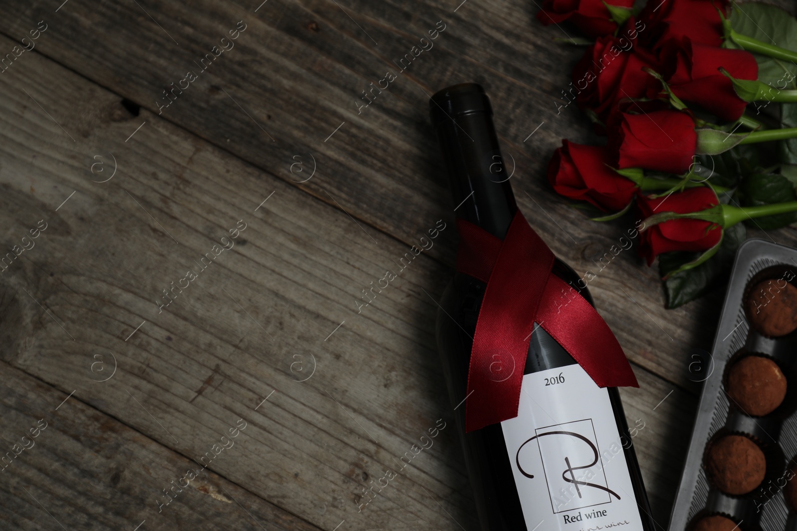 Photo of Chocolate sweets, bottle of red wine and roses on wooden table, flat lay. Space for text