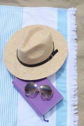 Beach towel with book, sunglasses and straw hat on sand, flat lay