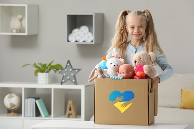Image of Humanitarian aid for Ukrainian refugees. Cute little girl holding donation box with toys indoors