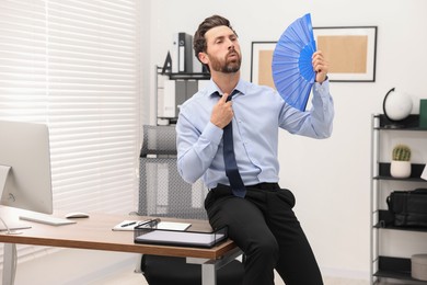 Bearded businessman waving blue hand fan to cool himself in office