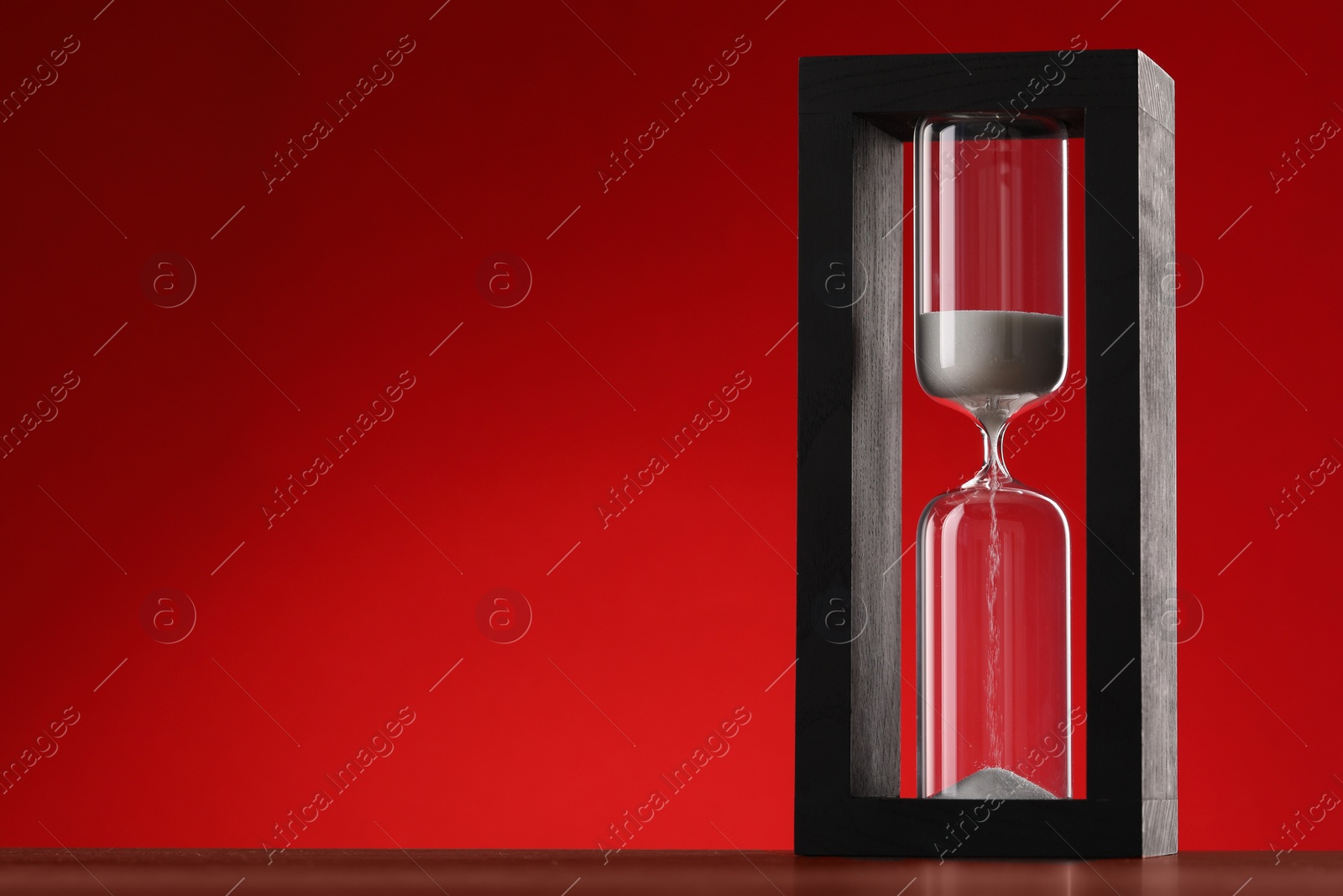 Photo of Hourglass with flowing sand on wooden table against red background, space for text