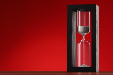 Hourglass with flowing sand on wooden table against red background, space for text