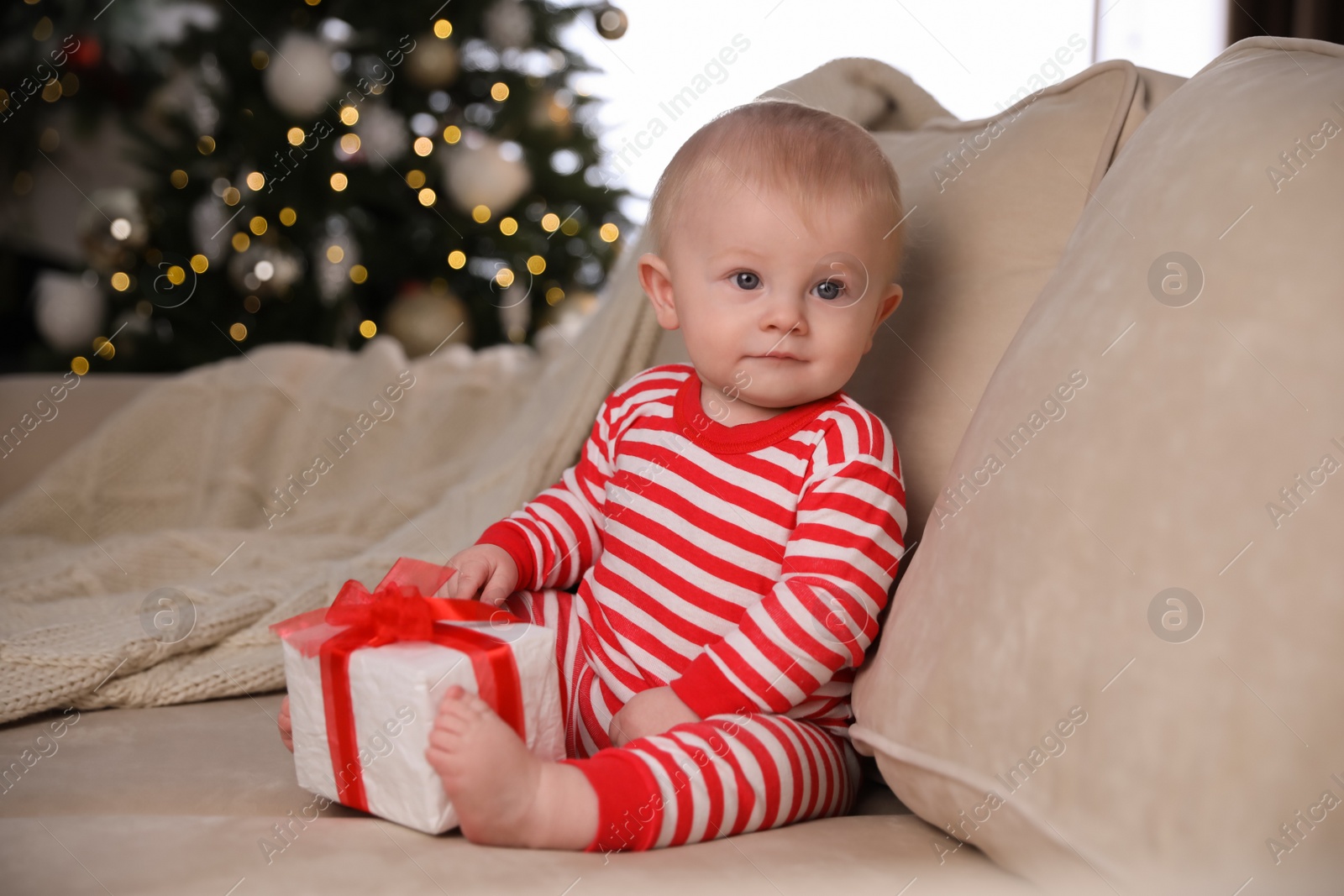 Photo of Cute baby in bright Christmas pajamas holding gift box on sofa at home