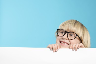 Cute little boy in glasses with blank board on light blue background, space for text