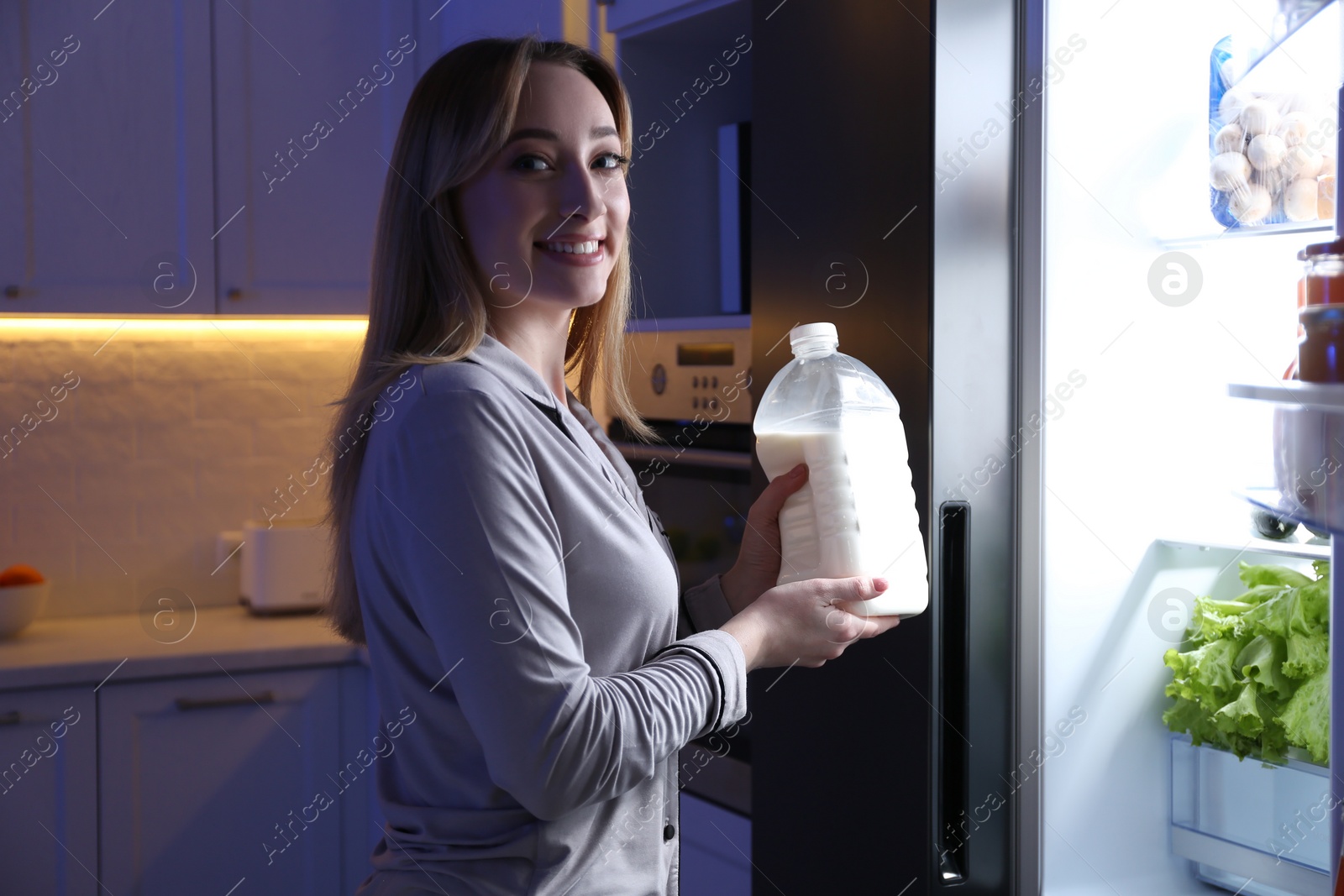 Photo of Young woman holding gallon bottle of milk near refrigerator in kitchen at night