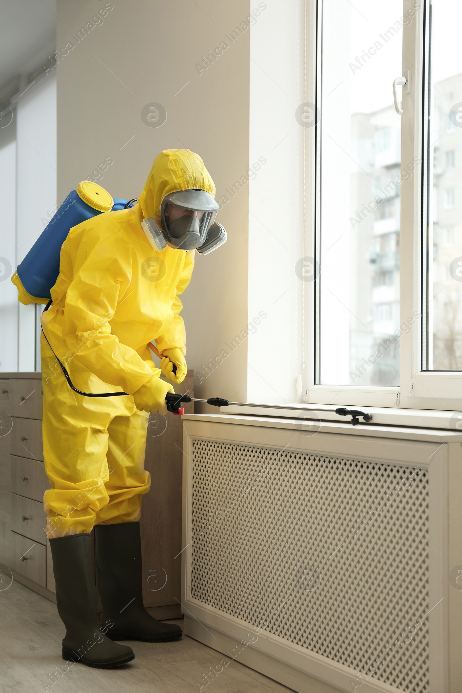 Photo of Pest control worker in protective suit spraying pesticide near window indoors