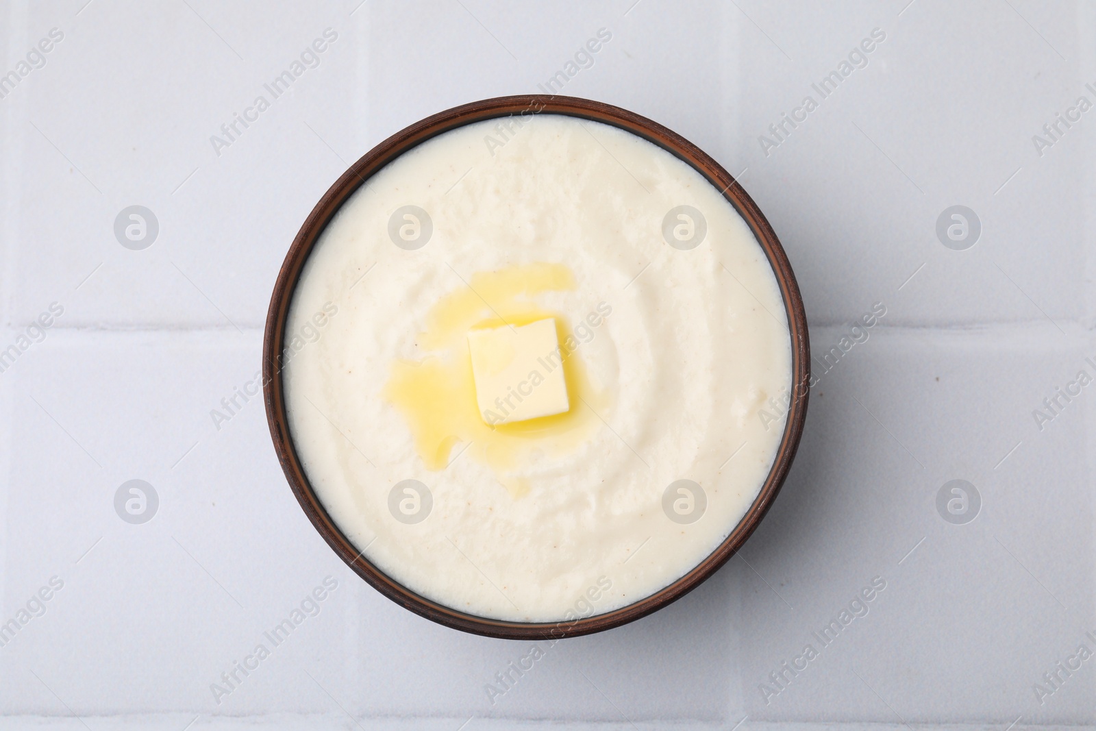 Photo of Delicious semolina pudding with butter in bowl on white tiled table, top view
