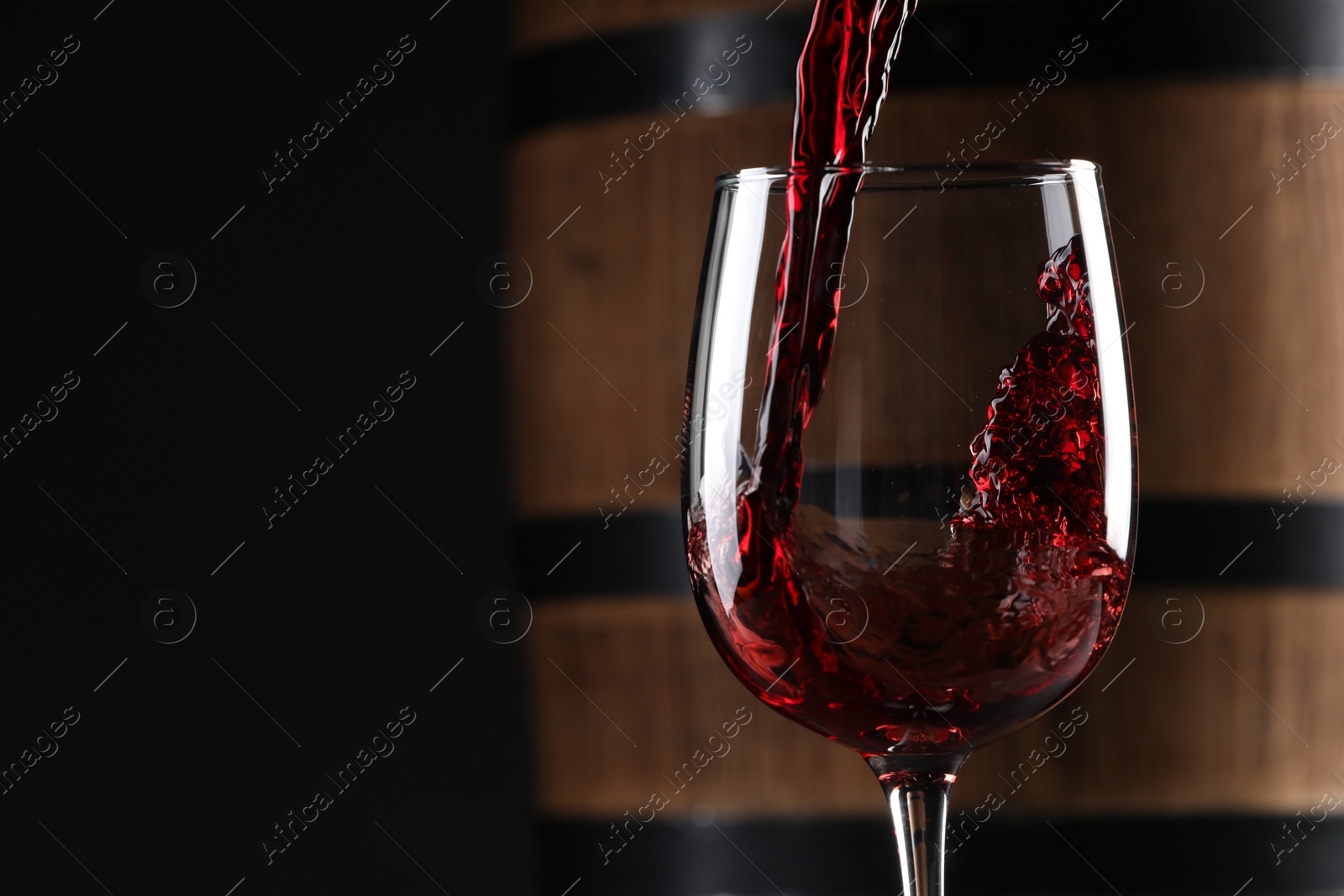 Photo of Pouring red wine into glass near wooden barrel against black background, closeup. Space for text