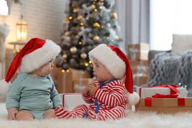 Image of Cute children in Santa hats on floor in room with Christmas tree