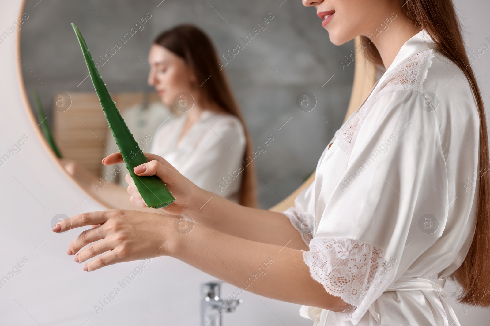 Photo of Young woman applying aloe gel from leaf onto her hand near mirror in bathroom, closeup