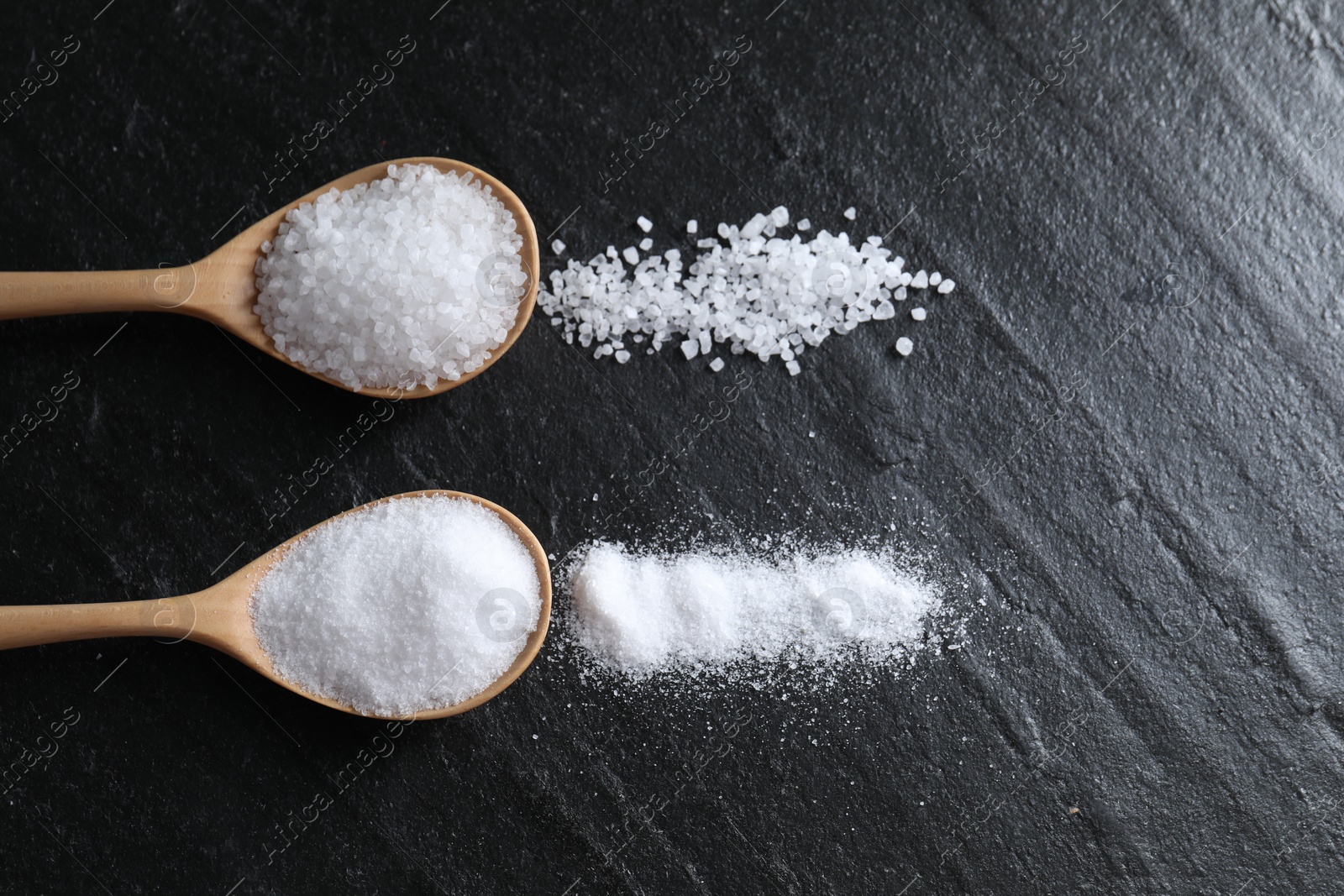 Photo of Organic salt in spoons on black table, flat lay. Space for text