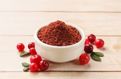 Dried cranberry powder in bowl, fresh berries and green leaves on light wooden table, closeup