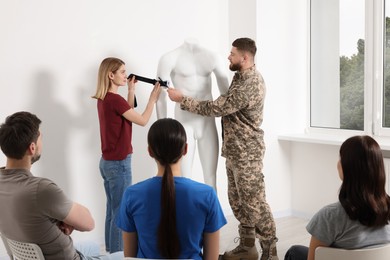 Photo of Soldier in military uniform teaching group of people how to apply medical tourniquet on mannequin indoors