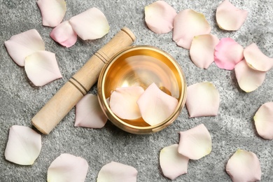 Golden singing bowl with petals and mallet on grey table, flat lay. Sound healing