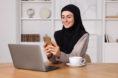 Muslim woman using smartphone near laptop at wooden table in room