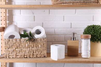 Photo of Toilet paper rolls in wicker basket, floral decor, cotton pads and dispenser on wooden shelf against white brick wall