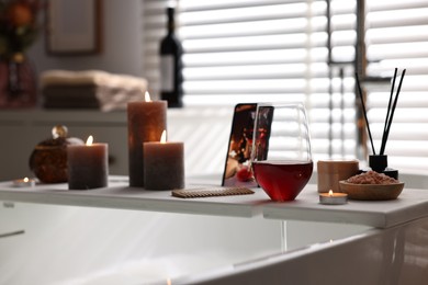 Photo of White wooden tray with smartphone, glass of wine and burning candles on bathtub in bathroom