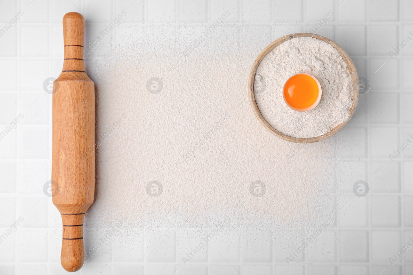 Photo of Flour, rolling pin and raw egg on white tiled table, top view