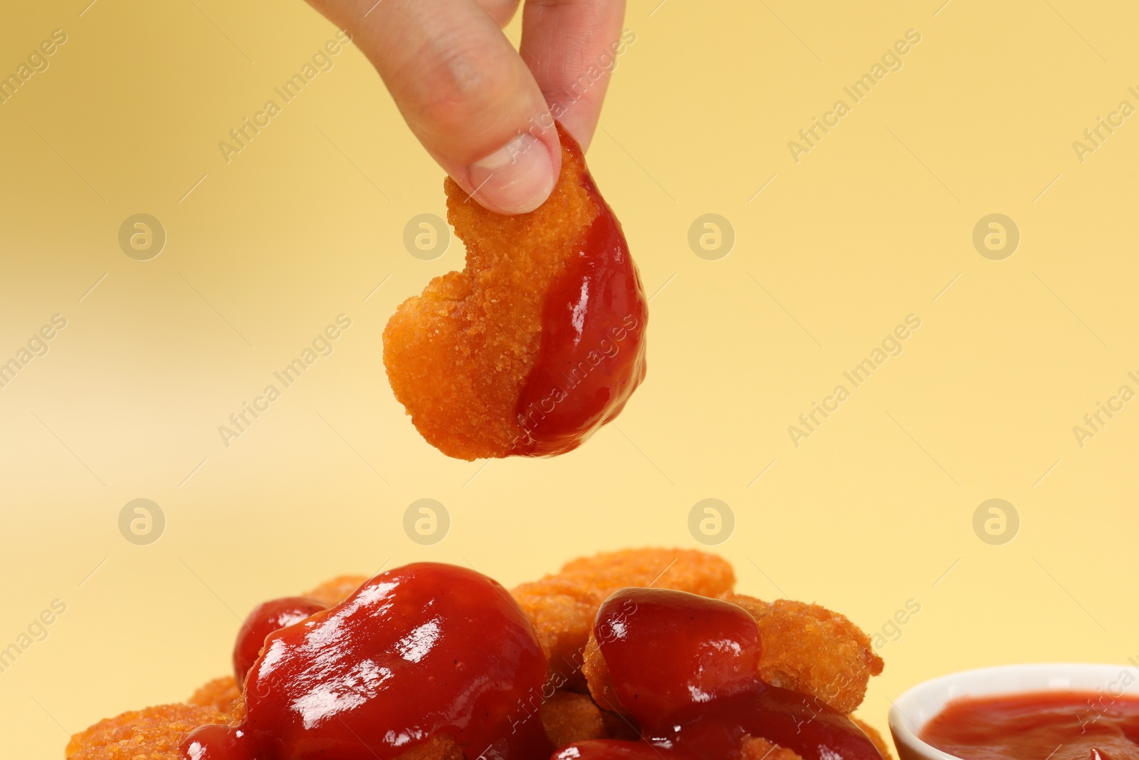 Photo of Woman holding delicious chicken nugget with ketchup on pale yellow background, closeup