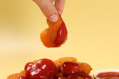 Woman holding delicious chicken nugget with ketchup on pale yellow background, closeup