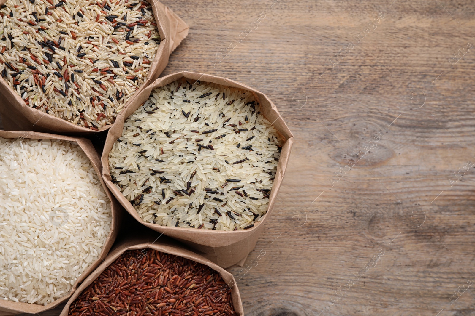 Photo of Different types of brown and polished rice in paper bags on wooden table, flat lay. Space for text