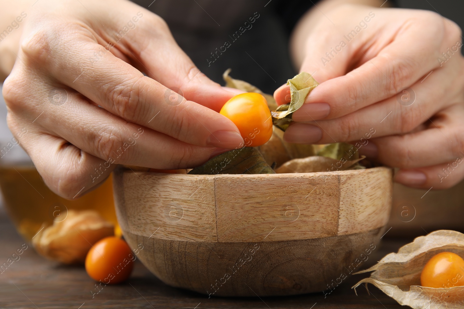 Photo of Woman peeling physalis fruit from calyxes at wooden table, closeup