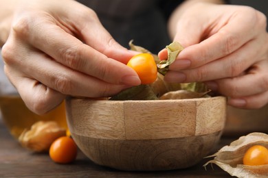 Photo of Woman peeling physalis fruit from calyxes at wooden table, closeup