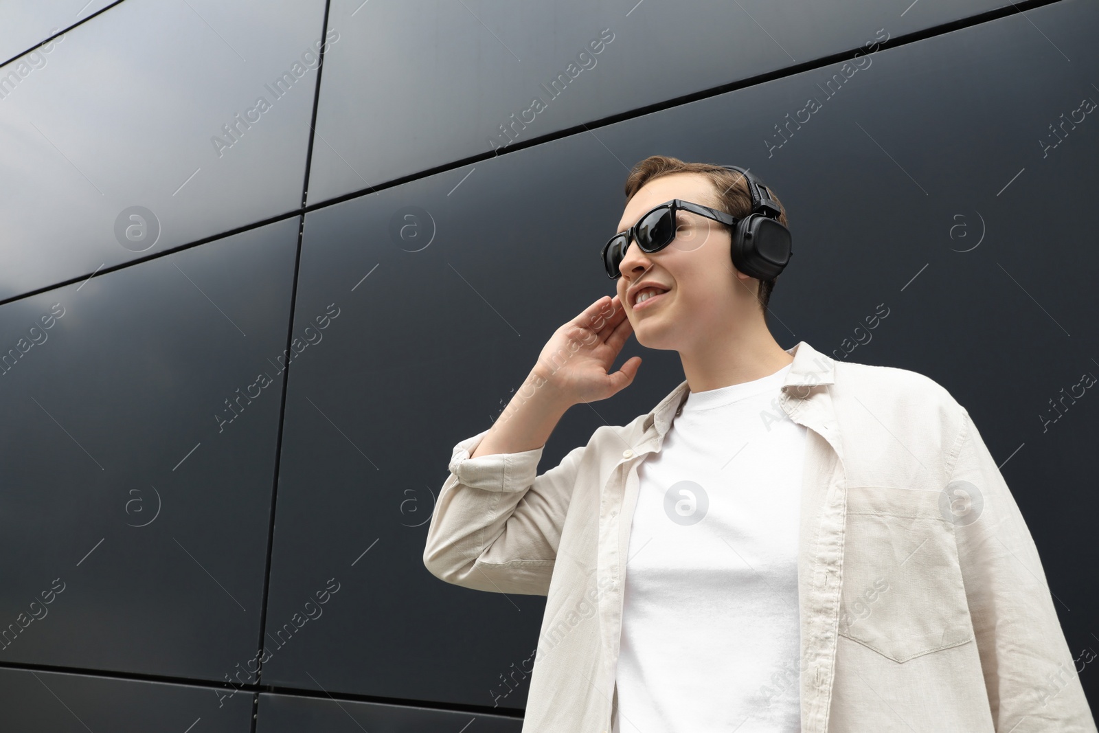 Photo of Handsome young man with headphones near black wall, low angle view. Space for text
