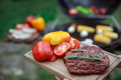 Photo of Woman holding wooden board with grilled vegetables and steak outdoors, closeup. Summer barbecue